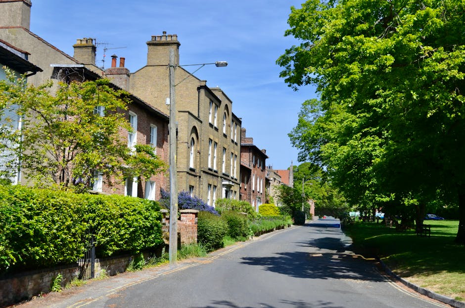 A quiet residential street with a paved tarmac road lined by well-maintained gardens and tall hedges on both sides. The left side features a sidewalk with a narrow strip of grass and a lamppost castin